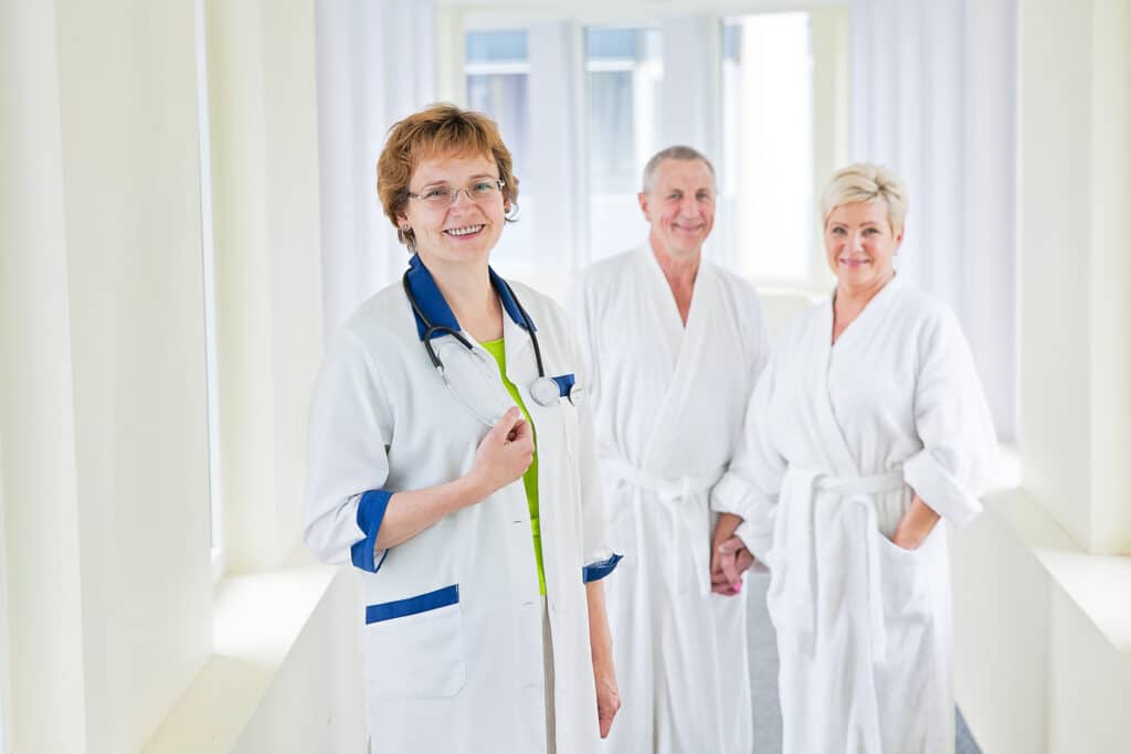 Гости лечебного центра курорта Вярска стоят вместе с врачом в галерее, соединяющей гостиницу санатория и лечебный корпус.